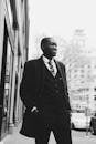 Low angle of young cheerful African American man in elegant suit and coat standing on city street keeping hand in pocket and looking away