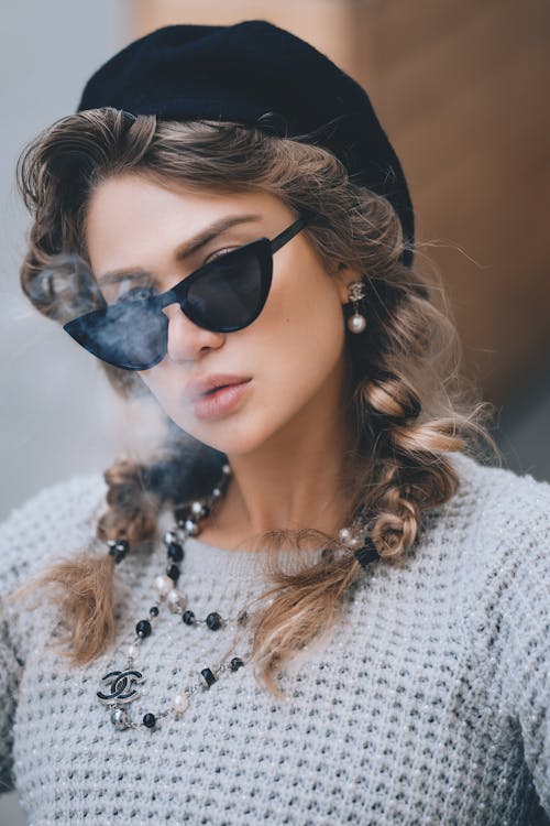 Young elegant woman with stylish sunglasses beret and accessories exhaling smoke looking at camera
