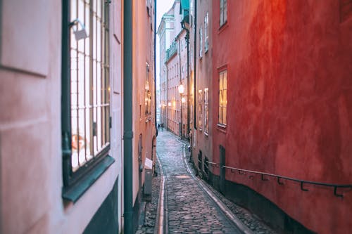 Narrow cobblestone pedestrian passage between old apartment buildings with burning electric lights in windows at twilight