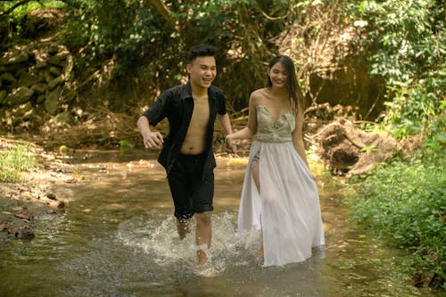 Cheerful ethnic couple holding hands while running towards camera in splashing water in sunny forest