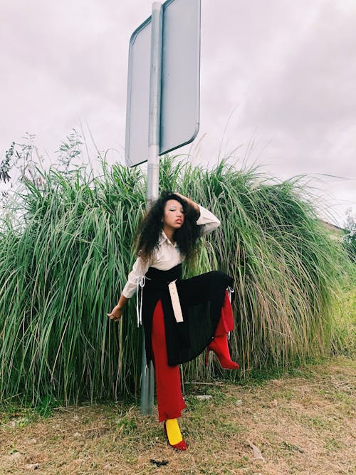 Confident Asian female with long curly hair touching hair and standing with raised leg near road sign against grassy bush in daytime