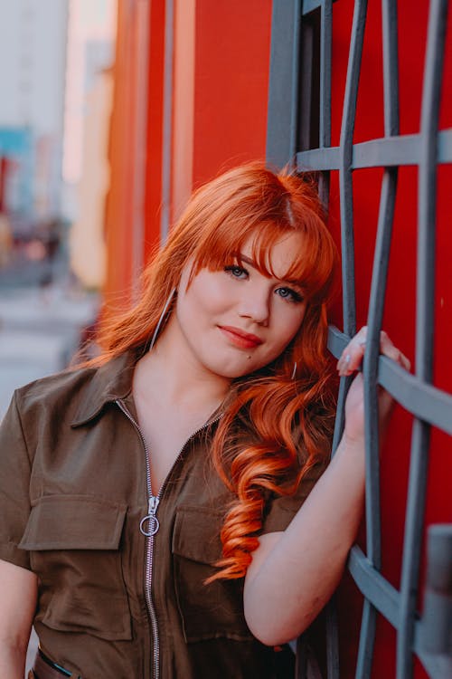 Free Positive young female with red hair leaning on metal fence and looking at camera while resting on street on sunny day Stock Photo