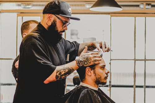 Side view of young tattooed bearded male hairdresser in casual clothes and eyeglasses cutting hair of client in modern barbershop