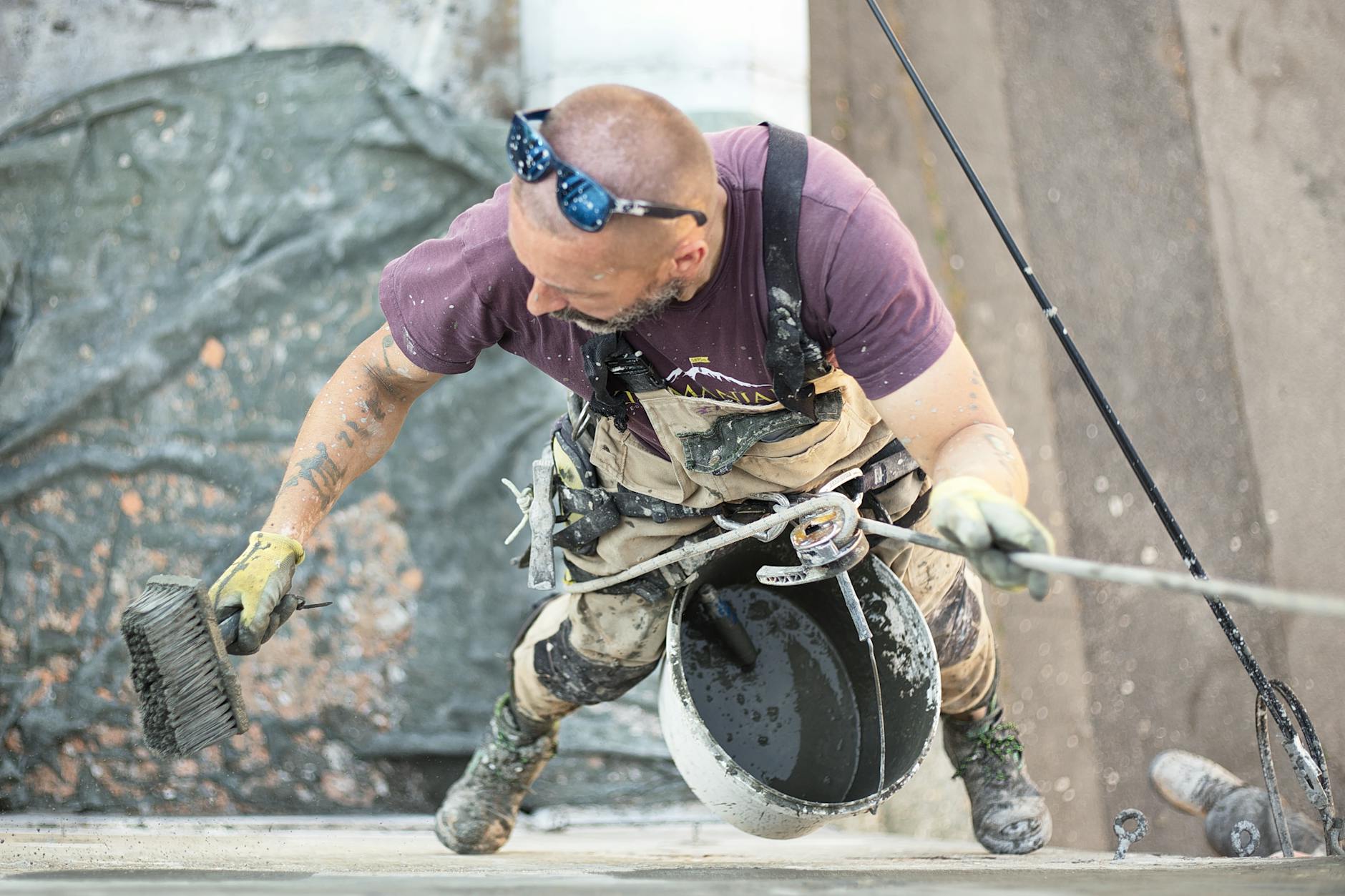 From above of man in safety harness hanging above ground and smoothing wall skim coat with liquid plaster using coarse brush