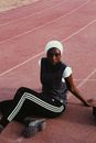 Full body of young pensive African American female runner in sportswear resting on dark red track in daytime
