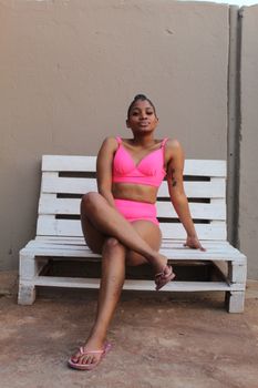 A woman sitting on a wooden bench in pink swimwear, exuding confidence in an outdoor setting.