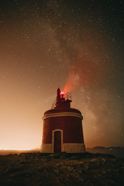 Low angle of red light glowing on tower of lighthouse against night sky with milky way and stars in twilight