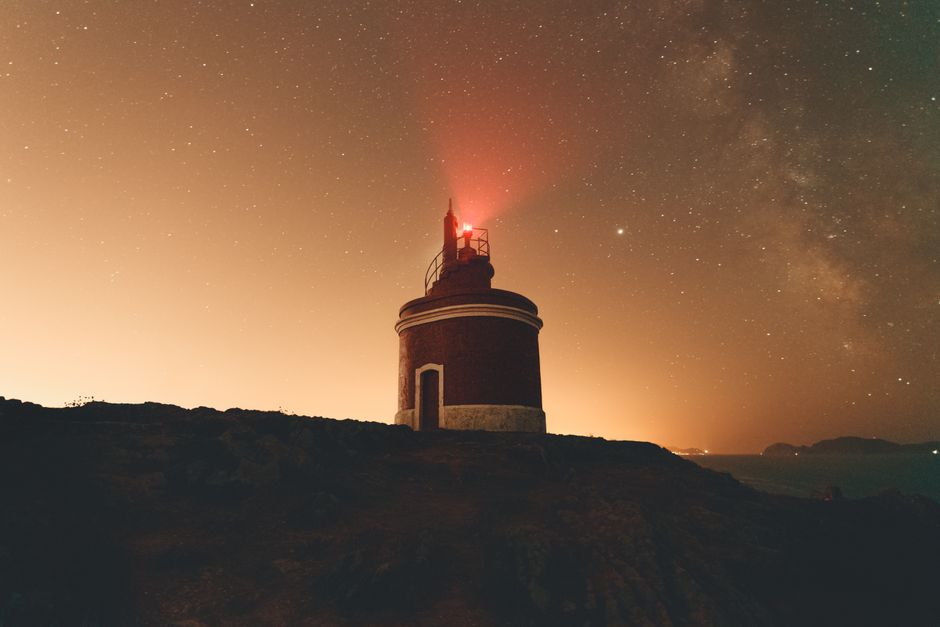 Low angle of glowing lighthouse with red beacon located on top of hill against night sky with bright stars in dark