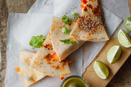 Close-up of tasty Mexican quesadilla with limes on a wooden board, garnished with herbs.