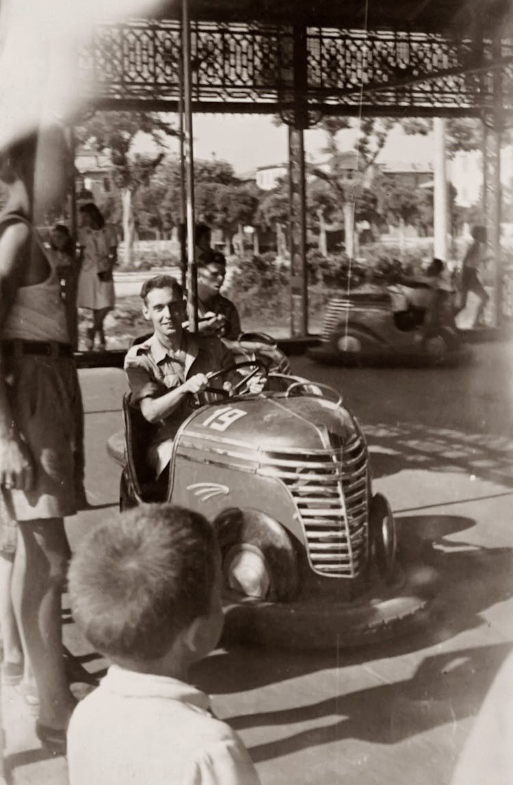 Grayscale Photo Of Man Having Fun Riding An Antique Car In The Fairground