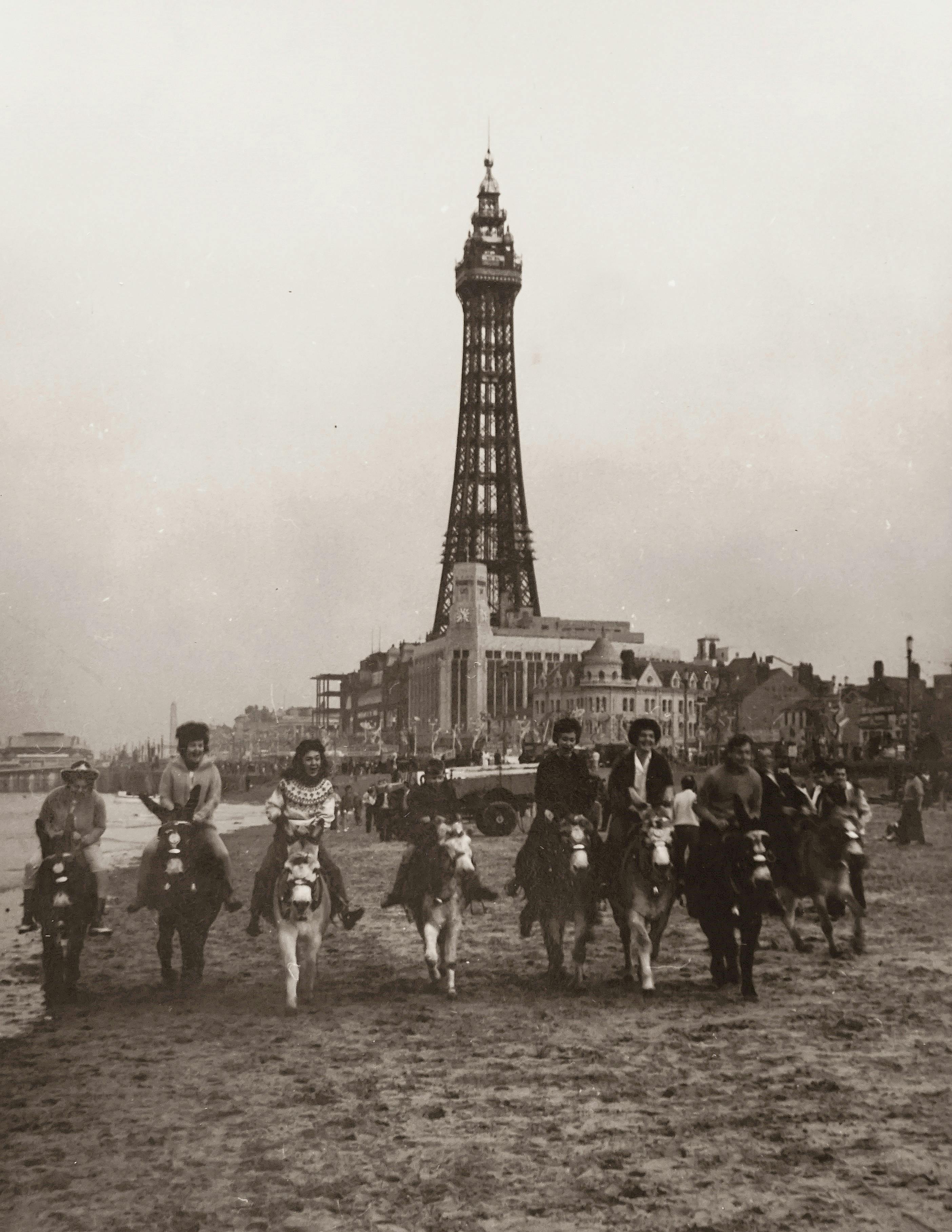 Historic group photo of people riding donkeys on the beach with Blackpool Tower in the background, classic style.