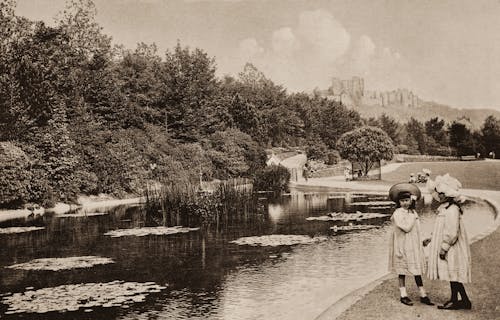 Grayscale Photo of Two Young Girls in White Dress Shirt Standing Near A Pond