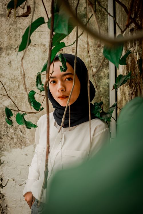 Free Portrait of a woman in a hijab standing by vines against a textured wall, wearing white long sleeves. Stock Photo