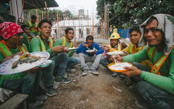 A group of construction workers in work attire enjoy a lunch break at an outdoor site.