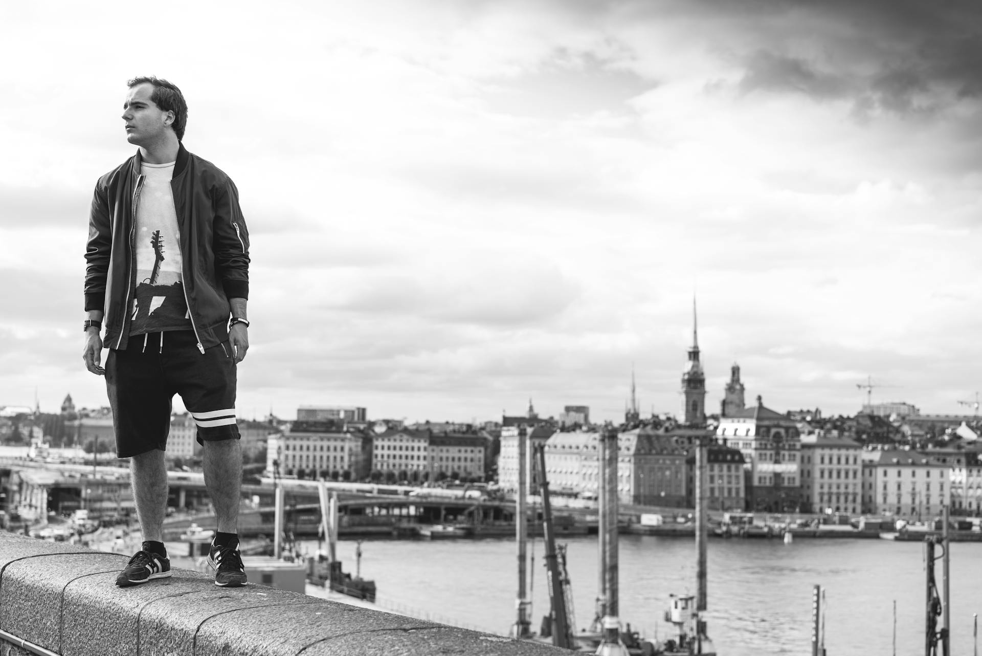 Urban scene of a young man standing on a bridge with Stockholm cityscape in the background.