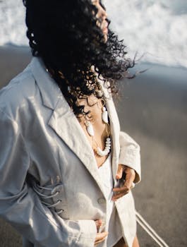 Stylish woman with curly hair wearing a white blazer and necklace by the ocean.