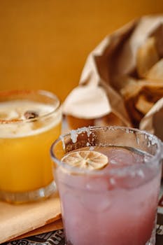 Two vibrant cocktails with lime garnishes alongside a bag of crispy chips.