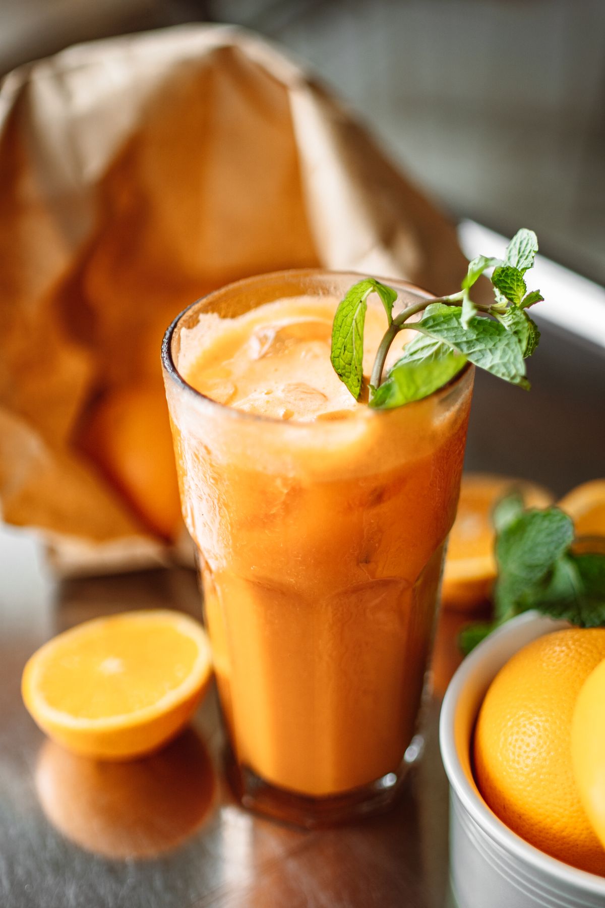 Tall glass of carrot orange juice with ice and a mint sprig beside halved oranges