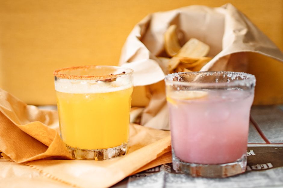 Close-up of vibrant citrus cocktails with salt rims and a bag of chips on paper.