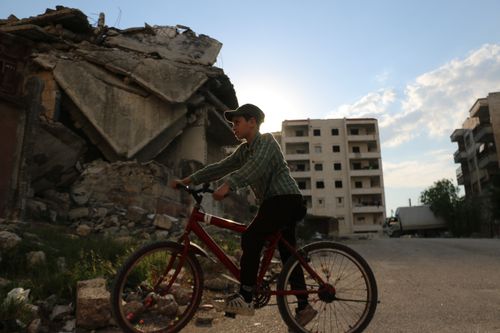 Free Side view of preteen in cap riding bicycle on paved road near destroyed building against cloudy sky in city in daylight Stock Photo