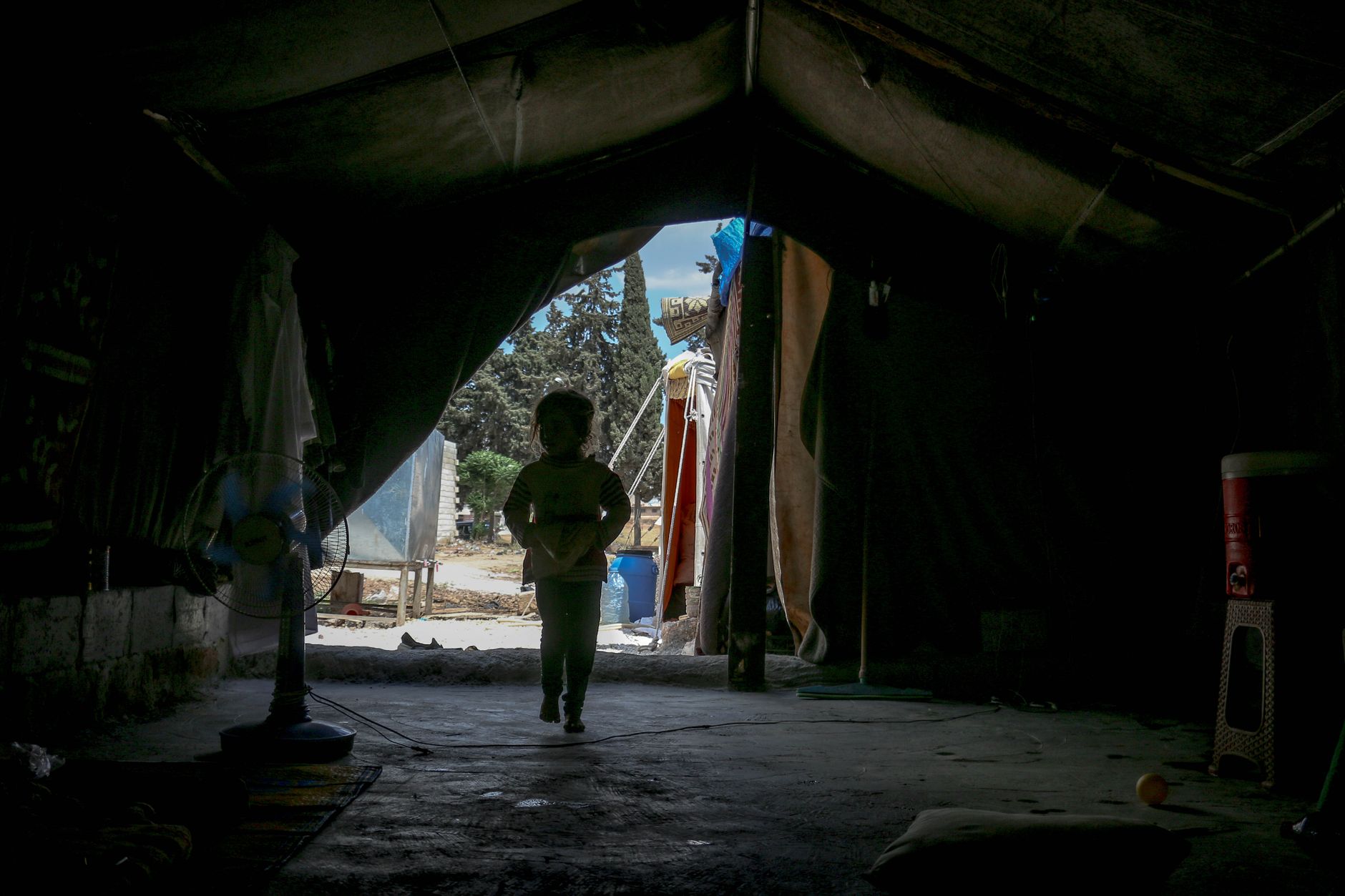 Silhouette of anonymous little kid walking in dark tent under roof in daytime in poor neighborhood with green trees in distance
