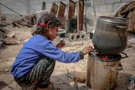 Side view of ethnic kid sitting near furnace with metal pan placed on fire in daylight in poor destroyed district