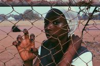 Black thoughtful woman wearing white turban standing behind wire fence on sports ground looking at camera