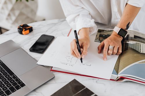 Free Woman in modern office writing on paper with laptop and smartphone nearby, focusing on design planning. Stock Photo