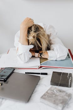 Overwhelmed woman resting head on desk with laptop and papers in home office.