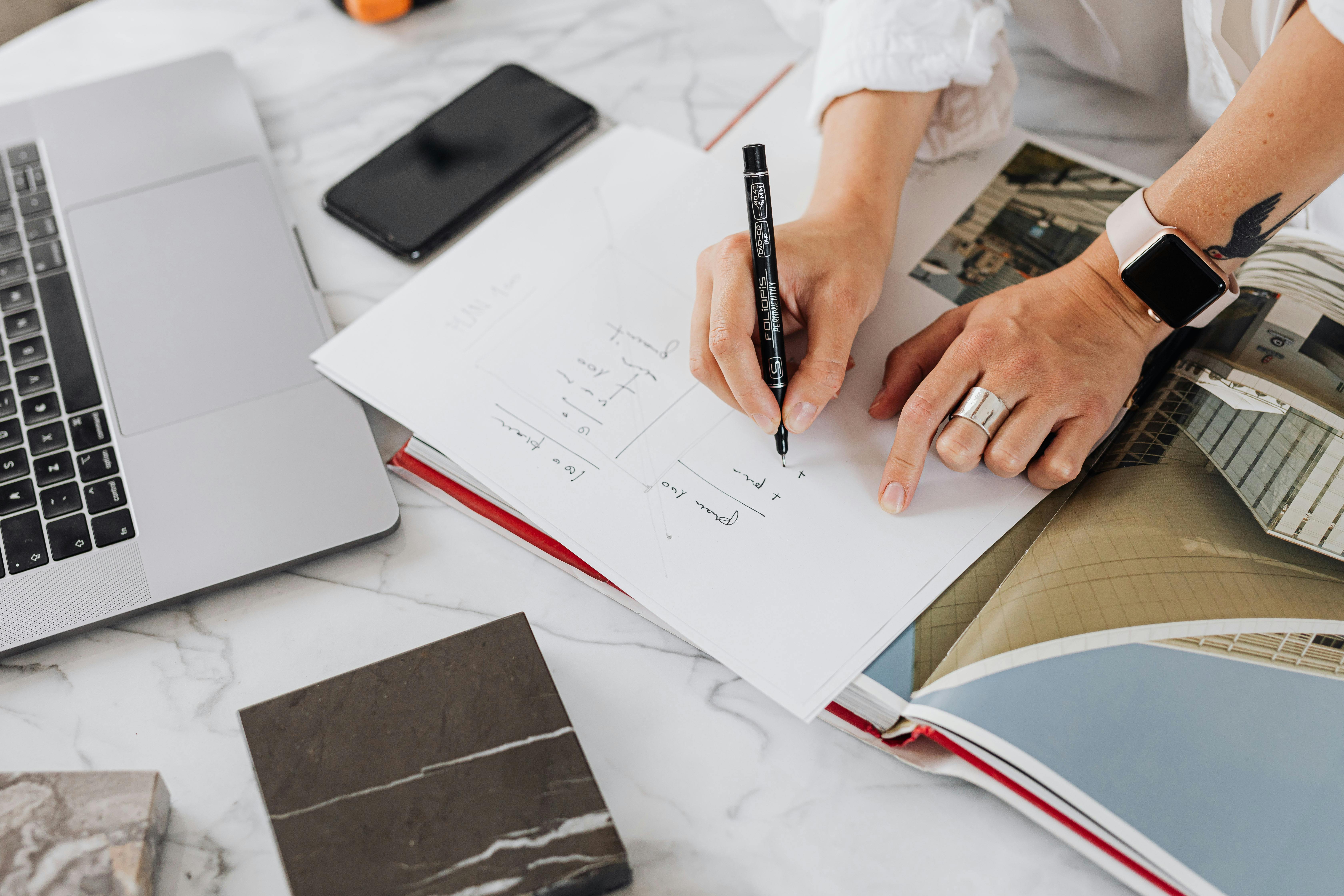 Hands writing on paper at a desk with laptop and smartphone in a modern office setting.
