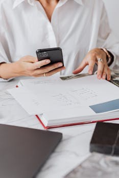 Close-up of a person holding a smartphone while reviewing diagrams on paper, showcasing multitasking in a modern work setting.