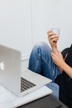 一名穿着牛仔裤、拿着白色马克杯的女子正坐着使用笔记本电脑
