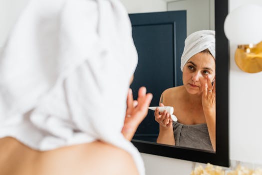 Woman applying facial moisturizer in front of a mirror with a towel wrap in a modern bathroom setting.