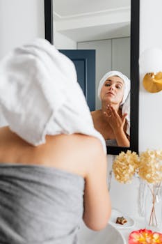 A woman with a towel on her head applying skincare cream in a bathroom mirror reflection.