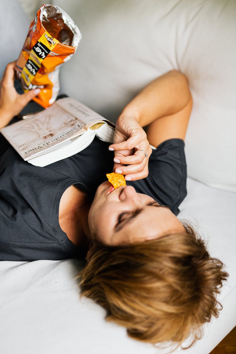 Woman In Gray Shirt Lying On Bed Eating Chips