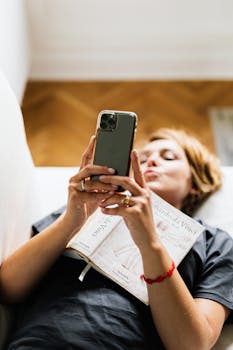A woman lying on a bed, using a smartphone while holding a book, enjoying a relaxed indoor moment.