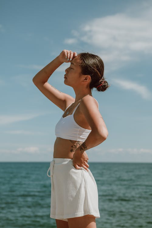 Side view of calm young female with eyes closed in white top and shorts holding hand on waist while standing on shore near sea