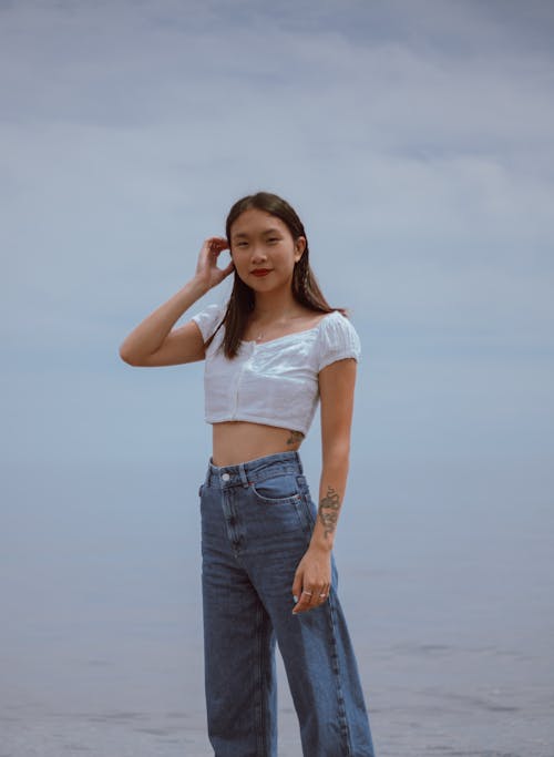 Young ethnic positive woman with tattoos standing on seashore near calm sea and smiling at camera