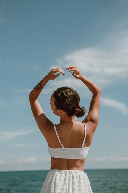 A woman raises her arms freely under the bright summer sky by the ocean.