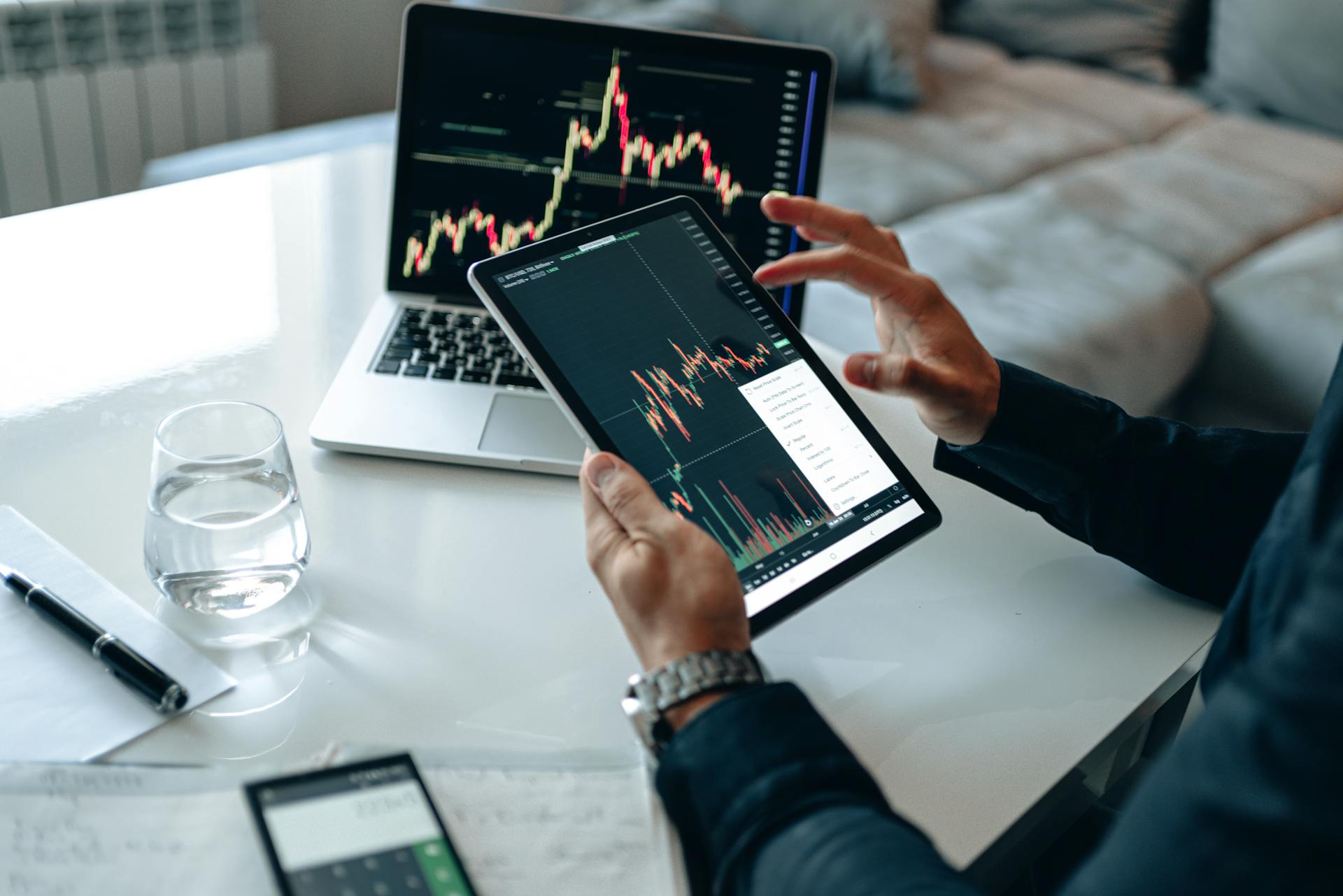 A Person in Blue Long Sleeves Holding a Tablet with Cryptocurrency Graph Neat Laptop on the Table