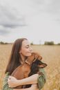 Happy young female with dark hair hugging adorable Dachshund dogs with closed eyes while spending time in wheat field in countryside