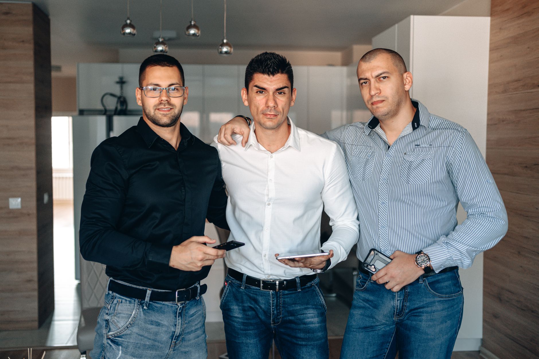 Three men in casual-formal attire standing in a modern office holding digital devices.