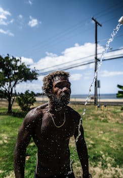 African American male with muscular torso standing on green field under pouring water with power line on background in summer day