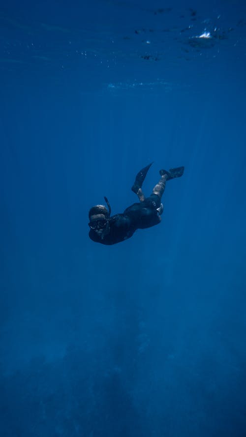 Unrecognizable diver in wetsuit and mask with tube swimming underwater in deep ocean with bubbles on surface in tropical country