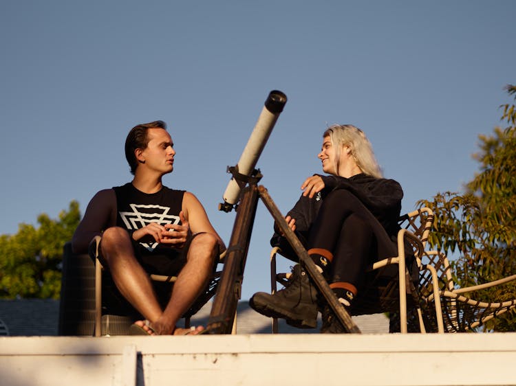 A Man And A Woman In Black Clothes Sitting  Outdoors Near A Telescope