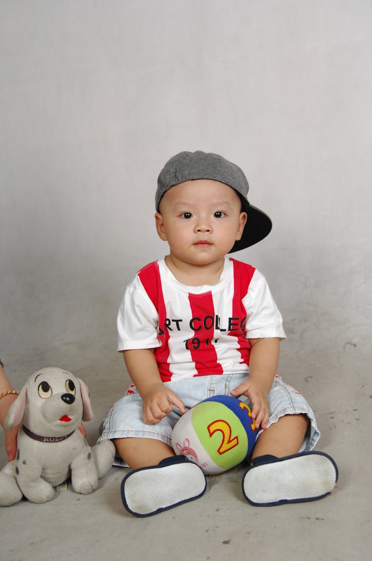 Toddler Wearing Cap Sitting While Holding A Plush Ball