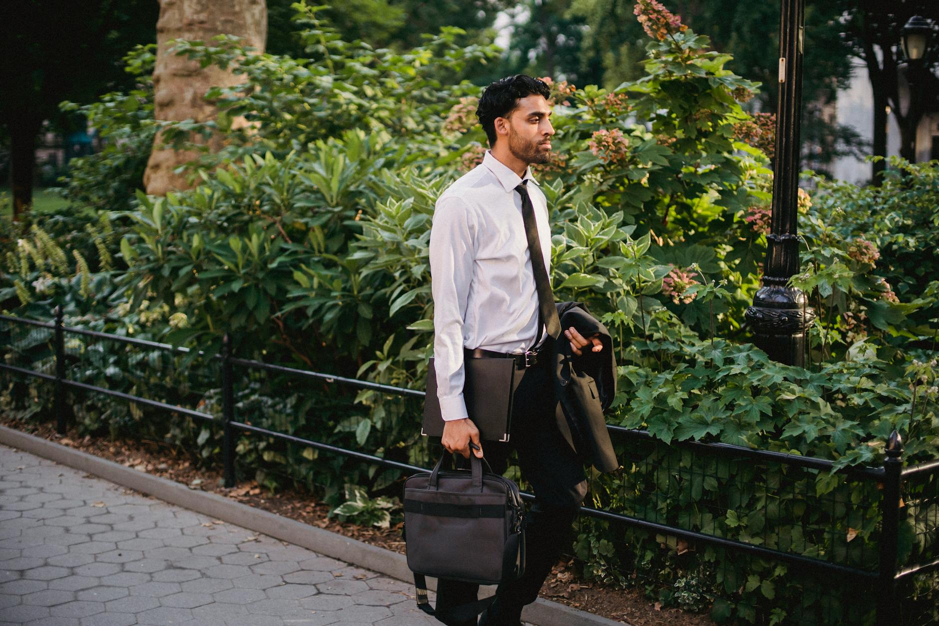 Businessman in formal attire carrying a briefcase, walking through a lush urban park on a sunny day.