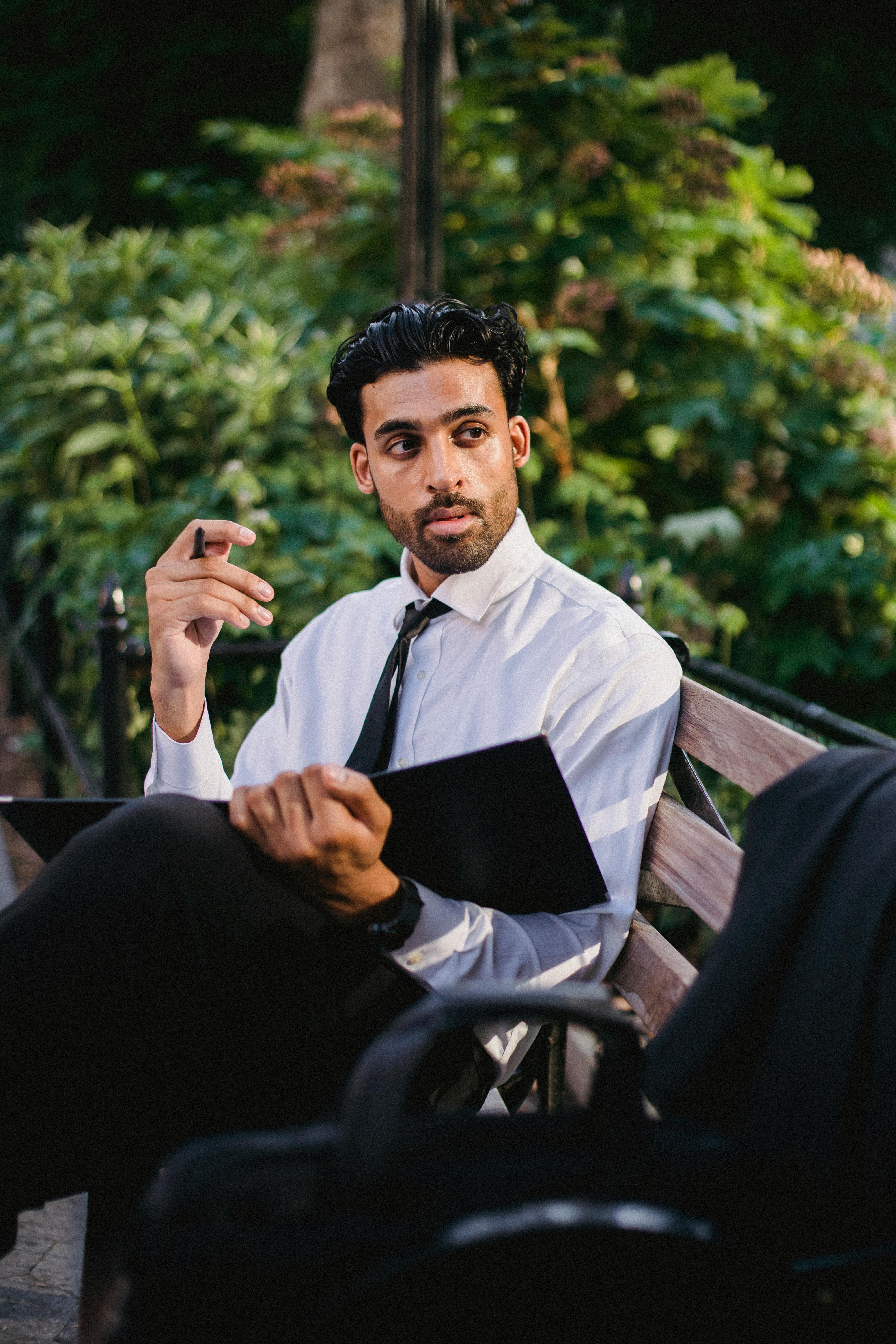 一位穿着正式的年轻专业人士坐在公园长凳上，手拿笔记本，陷入沉思