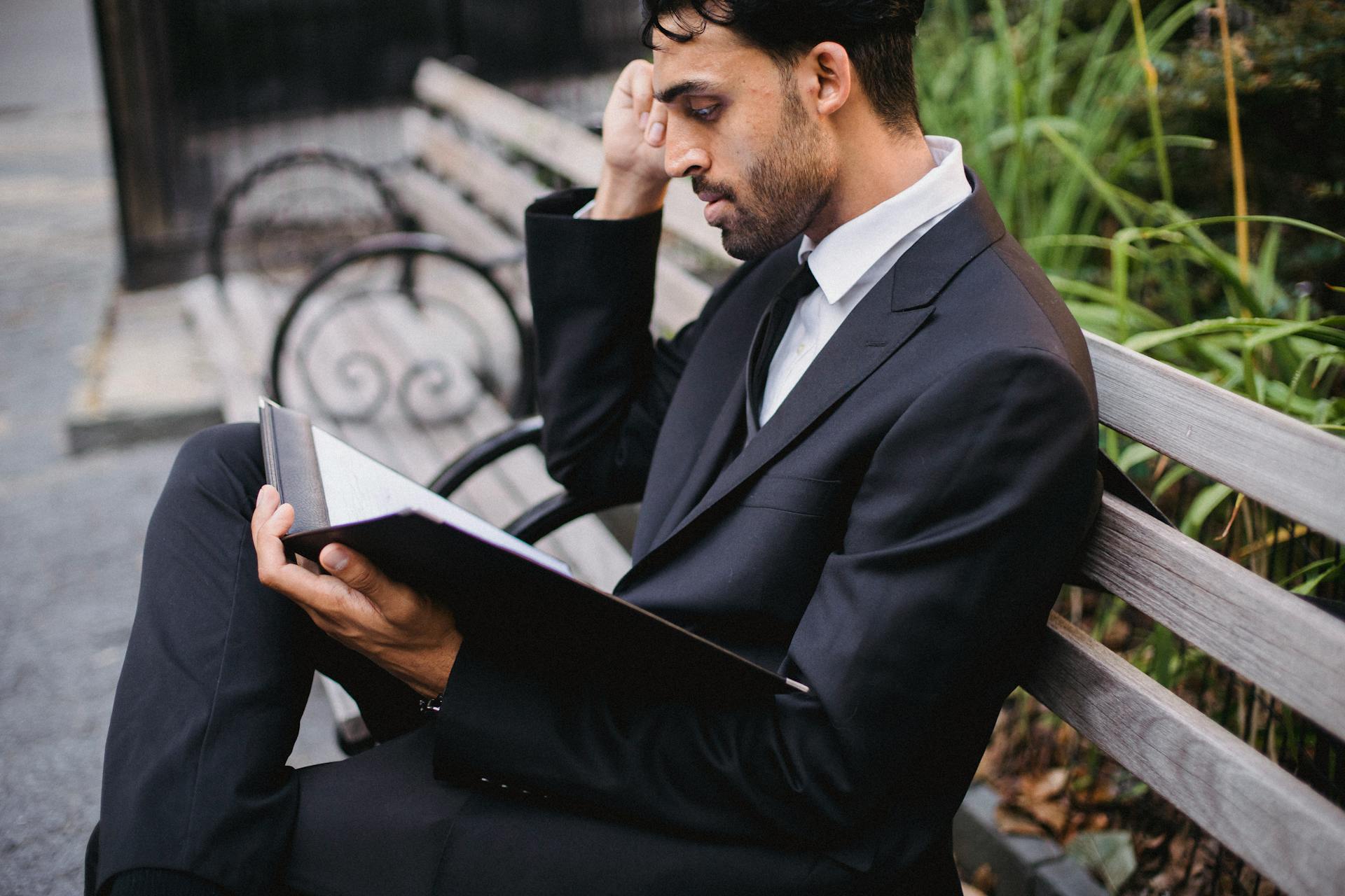 Man in a suit sitting on a park bench, focused on reading a folder. Outdoor setting, professional attire.