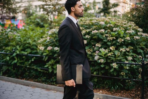 Free Side view of a well-dressed man walking through a city park holding a laptop. Stock Photo